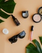 Various beauty and lifestyle items arranged aesthetically on a beige background. The items include a glass bottle with a dropper, an open jar of cream, a compact powder, a white candle, black sunglasses, a lip gloss, and large green leaves placed diagonally.