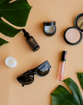 Various beauty and lifestyle items arranged aesthetically on a beige background. The items include a glass bottle with a dropper, an open jar of cream, a compact powder, a white candle, black sunglasses, a lip gloss, and large green leaves placed diagonally.