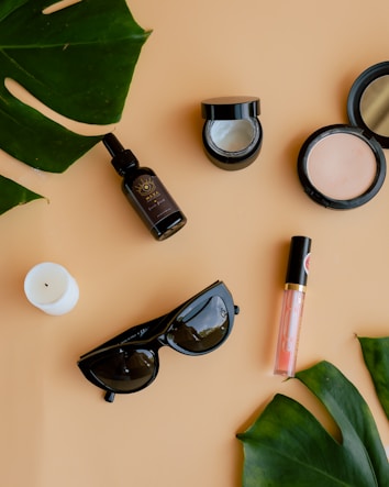 Various beauty and lifestyle items arranged aesthetically on a beige background. The items include a glass bottle with a dropper, an open jar of cream, a compact powder, a white candle, black sunglasses, a lip gloss, and large green leaves placed diagonally.