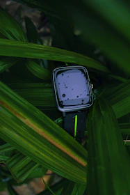 A modern smartwatch with a green strap displayed on a wrist against a leafy background.
