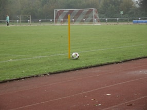A soccer field with a corner flag and a ball positioned next to it. A goalpost is visible in the background, and a player in light-colored attire stands on the field. The grass is lush and green, and the setting seems to be overcast.