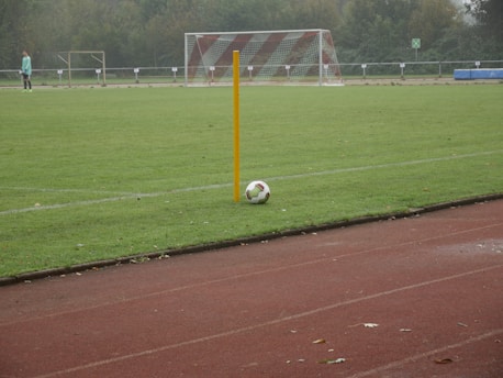 A soccer field with a corner flag and a ball positioned next to it. A goalpost is visible in the background, and a player in light-colored attire stands on the field. The grass is lush and green, and the setting seems to be overcast.