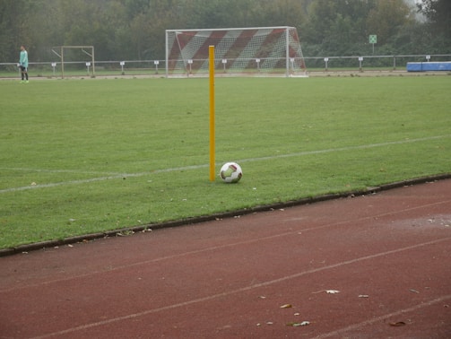 A soccer field with a corner flag and a ball positioned next to it. A goalpost is visible in the background, and a player in light-colored attire stands on the field. The grass is lush and green, and the setting seems to be overcast.