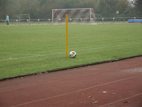 A soccer field with a corner flag and a ball positioned next to it. A goalpost is visible in the background, and a player in light-colored attire stands on the field. The grass is lush and green, and the setting seems to be overcast.