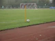 A soccer field with a corner flag and a ball positioned next to it. A goalpost is visible in the background, and a player in light-colored attire stands on the field. The grass is lush and green, and the setting seems to be overcast.