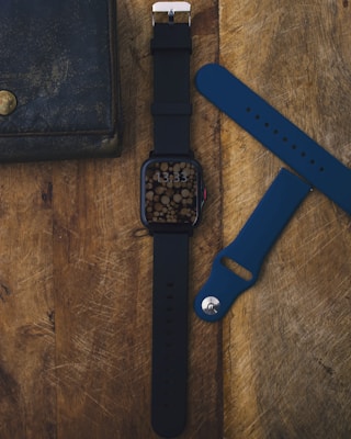 A smartwatch with a black strap is placed on a wooden surface. The watch face displays an artistic pattern with circular wooden textures. Alongside the watch, there is a detached blue watch strap and part of a dark, old leather wallet with a brass button visible.