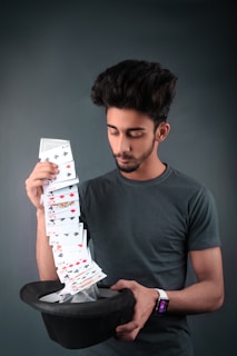 A young man with a neatly styled hairdo is skillfully holding a cascading stack of playing cards that appears to flow effortlessly into a black top hat. He wears a dark t-shirt and a smartwatch with a colorful display on his wrist. The background is a plain, muted color that does not distract from the main elements in the image.