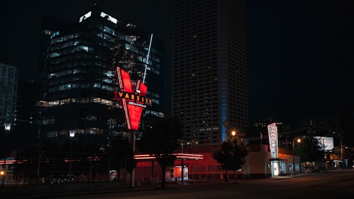 A city street scene at night featuring a prominently lit sign for 'The Varsity' with a bold red 'V'. Surrounding buildings are illuminated with soft, scattered lights from windows, and the street is empty with few visible streetlights casting a warm glow.