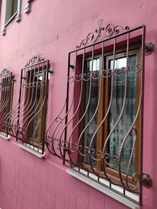 Ornate wrought iron bars cover three wooden windows set in a vibrant pink exterior wall, each window adorned with delicate lace curtains. The bars feature decorative scrollwork, adding an elegant touch to the otherwise simple architectural design.