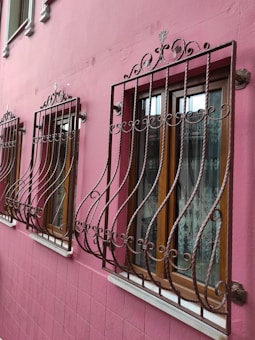 Ornate wrought iron bars cover three wooden windows set in a vibrant pink exterior wall, each window adorned with delicate lace curtains. The bars feature decorative scrollwork, adding an elegant touch to the otherwise simple architectural design.