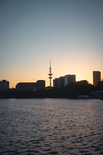 A serene city skyline at dusk with a prominent radio tower silhouetted against the fading light. Tall buildings line the horizon, and the calm water in the foreground reflects the twilight sky.