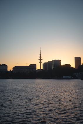 A serene city skyline at dusk with a prominent radio tower silhouetted against the fading light. Tall buildings line the horizon, and the calm water in the foreground reflects the twilight sky.