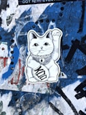 A black and white sticker of a waving cat with ornate designs and a coin with the word 'Kool' is pasted on a heavily graffitied and textured blue wall.