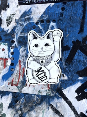 A black and white sticker of a waving cat with ornate designs and a coin with the word 'Kool' is pasted on a heavily graffitied and textured blue wall.