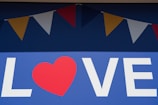 A large blue background displays the word 'LOVE' with a red heart replacing the letter 'O'. Above the text, a series of colorful triangular flags, alternating in red, yellow, and white, hang in a festive arrangement.