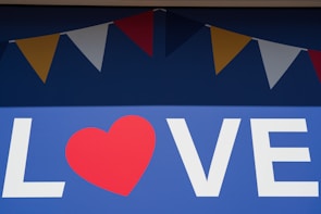 A large blue background displays the word 'LOVE' with a red heart replacing the letter 'O'. Above the text, a series of colorful triangular flags, alternating in red, yellow, and white, hang in a festive arrangement.