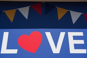A large blue background displays the word 'LOVE' with a red heart replacing the letter 'O'. Above the text, a series of colorful triangular flags, alternating in red, yellow, and white, hang in a festive arrangement.
