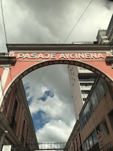 An archway labeled 'PASAJE AYCINENA' spans across a narrow street between two buildings. The sky is overcast with clouds, yet patches of blue are visible. The brickwork of the arch is painted in a muted pink color, framing the scene between historical architecture and a more modern high-rise building on the right.