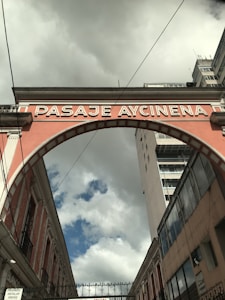 An archway labeled 'PASAJE AYCINENA' spans across a narrow street between two buildings. The sky is overcast with clouds, yet patches of blue are visible. The brickwork of the arch is painted in a muted pink color, framing the scene between historical architecture and a more modern high-rise building on the right.