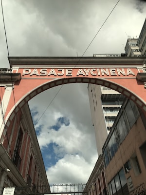 An archway labeled 'PASAJE AYCINENA' spans across a narrow street between two buildings. The sky is overcast with clouds, yet patches of blue are visible. The brickwork of the arch is painted in a muted pink color, framing the scene between historical architecture and a more modern high-rise building on the right.