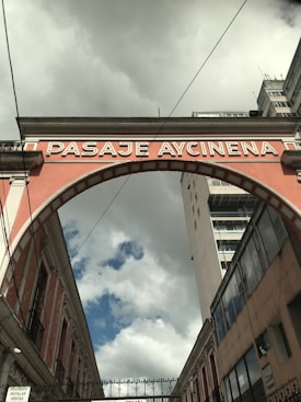 An archway labeled 'PASAJE AYCINENA' spans across a narrow street between two buildings. The sky is overcast with clouds, yet patches of blue are visible. The brickwork of the arch is painted in a muted pink color, framing the scene between historical architecture and a more modern high-rise building on the right.