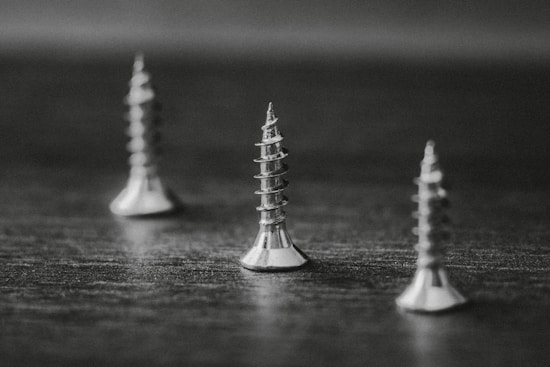 Three metal screws stand upright on a flat surface. The image is in black and white, creating a high contrast between the shiny screws and the dark background. The screws are evenly spaced with a focus on the one in the center, highlighting its spiral threads and pointed tip.