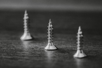 Three metal screws stand upright on a flat surface. The image is in black and white, creating a high contrast between the shiny screws and the dark background. The screws are evenly spaced with a focus on the one in the center, highlighting its spiral threads and pointed tip.
