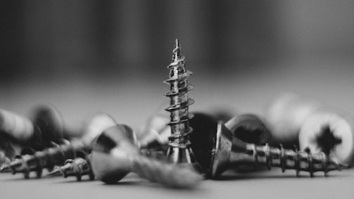 A collection of metallic screws scattered on a surface with one screw standing upright, highlighted against the out-of-focus background. The image is captured in black and white, emphasizing textures and shadows.