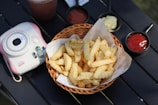 A wicker basket filled with crinkle-cut French fries is placed on a black surface. Accompanying the fries are small bowls of condiments, including ketchup and mayonnaise. To the side, there is an Instax mini camera with pink accents, and a beverage is partially visible.