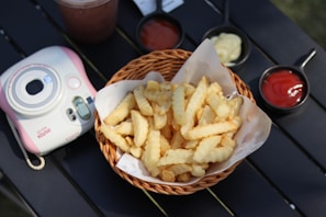 A wicker basket filled with crinkle-cut French fries is placed on a black surface. Accompanying the fries are small bowls of condiments, including ketchup and mayonnaise. To the side, there is an Instax mini camera with pink accents, and a beverage is partially visible.