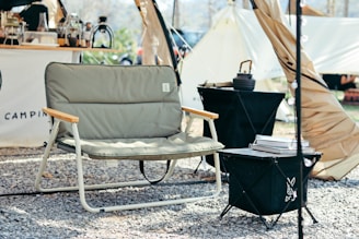 A relaxed campsite setting features a cushioned outdoor chair with wooden armrests positioned in front of a tent. Nearby, a small black camping table holds stacked books and a kettle. The ground is covered with gravel, and various camping supplies are visible in the background.