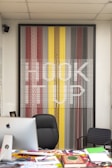 A modern office setup featuring a large, colorful artwork on the wall that spells out 'HOOK UP' against a pattern of vertical lines in shades of red, yellow, gray, and black. In front of the artwork, there is a black office chair, a desk cluttered with magazines and a computer, and a smaller patterned chair to the side.
