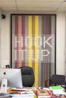 A modern office setup featuring a large, colorful artwork on the wall that spells out 'HOOK UP' against a pattern of vertical lines in shades of red, yellow, gray, and black. In front of the artwork, there is a black office chair, a desk cluttered with magazines and a computer, and a smaller patterned chair to the side.
