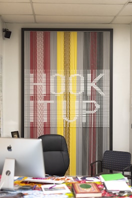 A modern office setup featuring a large, colorful artwork on the wall that spells out 'HOOK UP' against a pattern of vertical lines in shades of red, yellow, gray, and black. In front of the artwork, there is a black office chair, a desk cluttered with magazines and a computer, and a smaller patterned chair to the side.