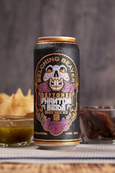 A tall can of craft beer is adorned with vibrant and intricate artwork featuring a colorful skull and floral designs. The label includes the text 'Belching Beaver,' 'Deftones,' and 'Phantom Bride.' Behind the can, there are snacks including a glass of green sauce and a stack of chips.