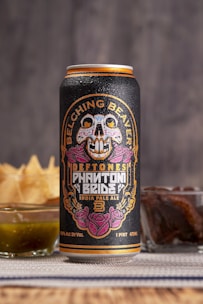 A tall can of craft beer is adorned with vibrant and intricate artwork featuring a colorful skull and floral designs. The label includes the text 'Belching Beaver,' 'Deftones,' and 'Phantom Bride.' Behind the can, there are snacks including a glass of green sauce and a stack of chips.