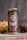 A tall can of craft beer is adorned with vibrant and intricate artwork featuring a colorful skull and floral designs. The label includes the text 'Belching Beaver,' 'Deftones,' and 'Phantom Bride.' Behind the can, there are snacks including a glass of green sauce and a stack of chips.