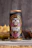 A tall can of craft beer is adorned with vibrant and intricate artwork featuring a colorful skull and floral designs. The label includes the text 'Belching Beaver,' 'Deftones,' and 'Phantom Bride.' Behind the can, there are snacks including a glass of green sauce and a stack of chips.