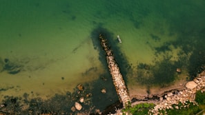 An aerial view of a coastal area featuring a stone breakwater extending into clear green waters. Various rocks are scattered around the shore, and a small boat is anchored near the breakwater. The surrounding area has patches of green vegetation.