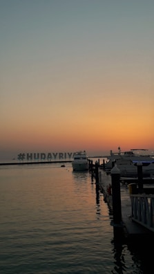 A serene marina during sunset, with calm waters reflecting the soft hues of the sky. Several boats and yachts are docked along a wooden pier, and the silhouette of a large hashtag sign is visible in the distance, spelling '#HUDAYRIYAT'. The scene is tranquil and captures the transition from day to night.
