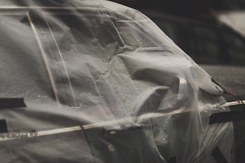 A close-up view of a car covered in a plastic sheet secured with tape. The plastic sheet is wet with visible water droplets, suggesting recent rain. There are other cars in the background, slightly out of focus.