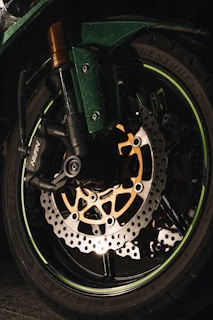 A close-up view of a motorcycle wheel, highlighting the intricate design of the brake disc and the surrounding components. The dark tones are contrasted by the illumination on the metallic parts, giving a detailed look at the engineering.