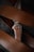 A detailed view of a folding knife with a metallic blade and a brown, possibly copper-toned handle. The knife is partially hidden behind out-of-focus brown elements, potentially leather straps or belts, creating a layered composition.