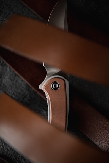 A detailed view of a folding knife with a metallic blade and a brown, possibly copper-toned handle. The knife is partially hidden behind out-of-focus brown elements, potentially leather straps or belts, creating a layered composition.
