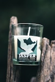 A glass candle with a label that reads 'Jasper National Park' is placed on a rustic wooden surface. The label features a mountain landscape design. The candle is cylindrical and white, emitting a clean, simple aesthetic.
