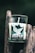 A glass candle with a label that reads 'Jasper National Park' is placed on a rustic wooden surface. The label features a mountain landscape design. The candle is cylindrical and white, emitting a clean, simple aesthetic.