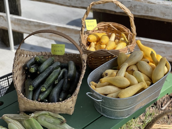 Baskets and a galvanized metal tub filled with fresh vegetables are displayed on a green table. The basket on the left contains green zucchinis, while the other two baskets and the metal tub contain yellow squashes. Corn is also partially visible in the foreground. Price tags are visible on two of the baskets.