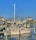 A marina with several docked motorboats, featuring a prominent beige boat named 'Seas the Day'. The sky is clear and blue, and the surrounding buildings are coastal houses painted in muted tones. The water reflects the boats and docks partially visible in the foreground.