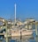A marina with several docked motorboats, featuring a prominent beige boat named 'Seas the Day'. The sky is clear and blue, and the surrounding buildings are coastal houses painted in muted tones. The water reflects the boats and docks partially visible in the foreground.