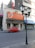 A red vintage car is parked on the street in front of a pharmacy with a large orange sign that reads 'Del Pueblo Farmacias'. The area appears urban with buildings in the background. A tall black lamppost stands near the curb.